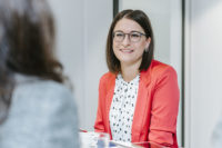 Frau in Blazer sitzt lächelnd am Tisch.