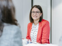 Woman in blazer is sitting at the table smiling.